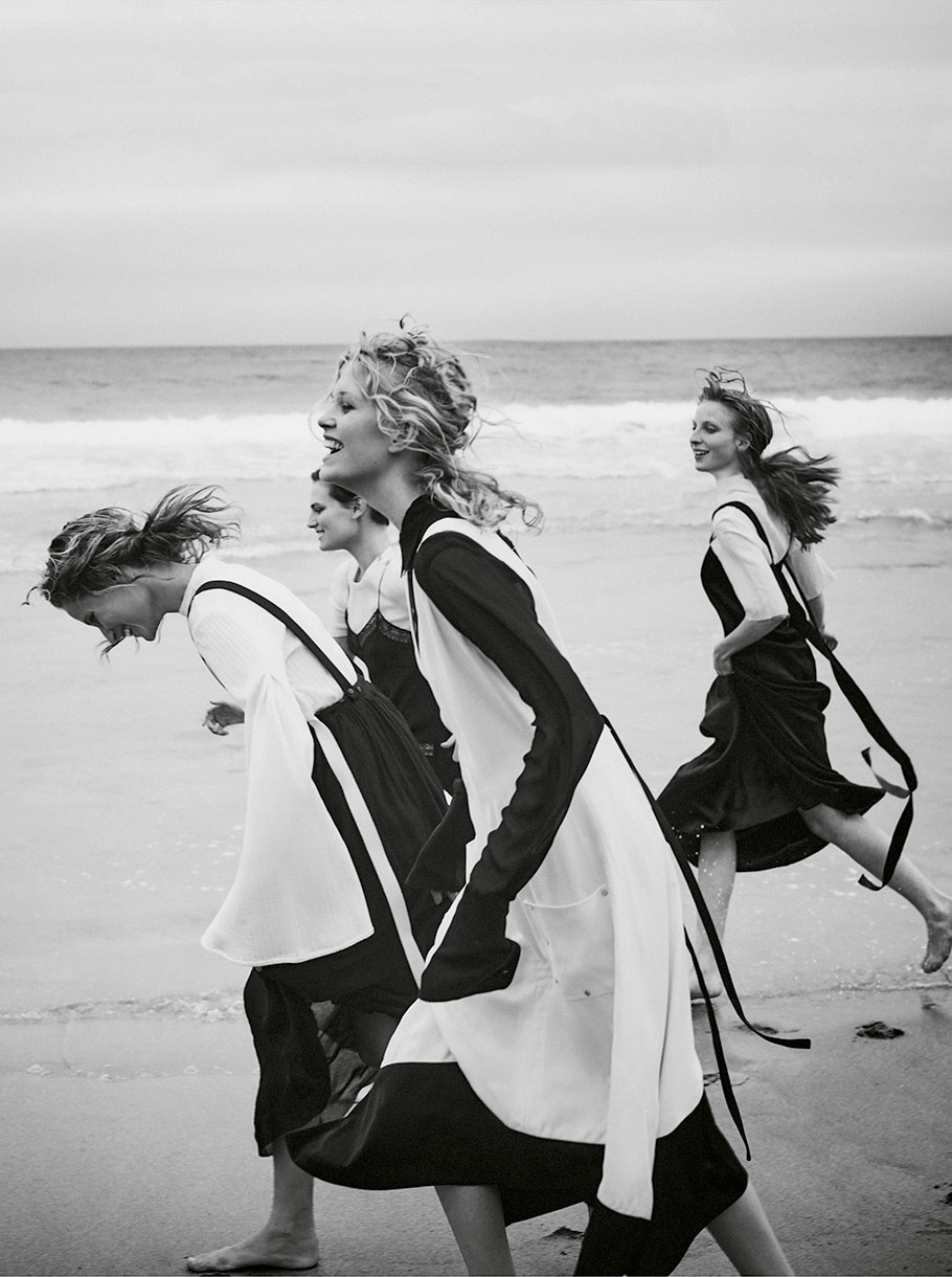 Four adult women in black and white dresses run barefoot on a sandy beach by the ocean under an overcast sky in a black and white fashion shot.