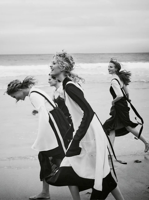 Four adult women in black and white dresses run barefoot on a sandy beach by the ocean under an overcast sky in a black and white fashion shot.
