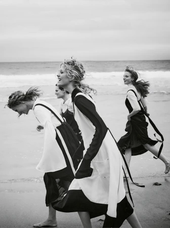 Four adult women in black and white dresses run barefoot on a sandy beach by the ocean under an overcast sky in a black and white fashion shot.