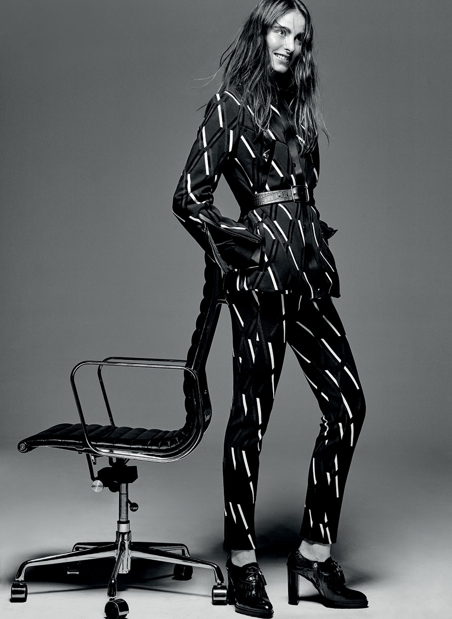A woman in a patterned black and white suit poses next to a black office chair in a fashion editorial shot against a gray background.