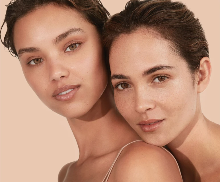 Close-up portrait of two young adult women with natural makeup, light skin, and brown hair posing against a light peach-colored background.