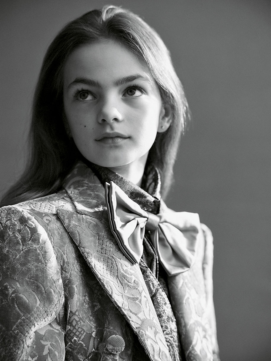 A young person with light hair, wearing a patterned blazer and bow blouse, looks upward in a black and white studio portrait.