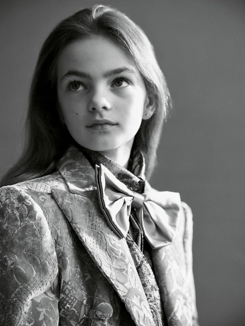 A young person with light hair, wearing a patterned blazer and bow blouse, looks upward in a black and white studio portrait.