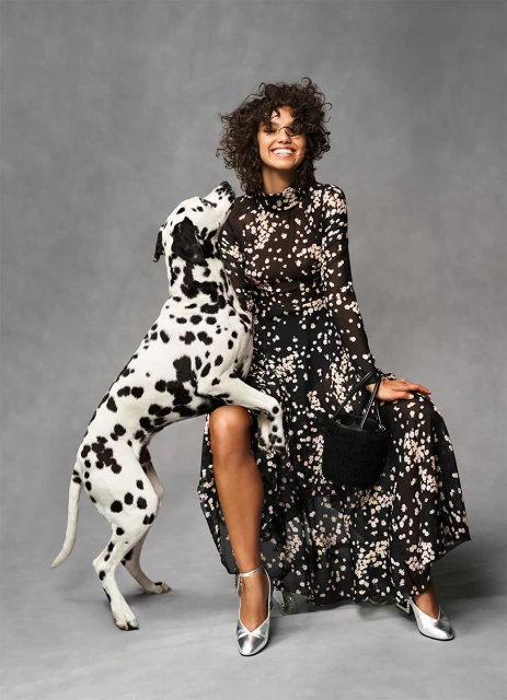 A curly-haired woman in a long floral dress sits next to a Dalmatian dog against a gray background in a fashion forward studio shot.