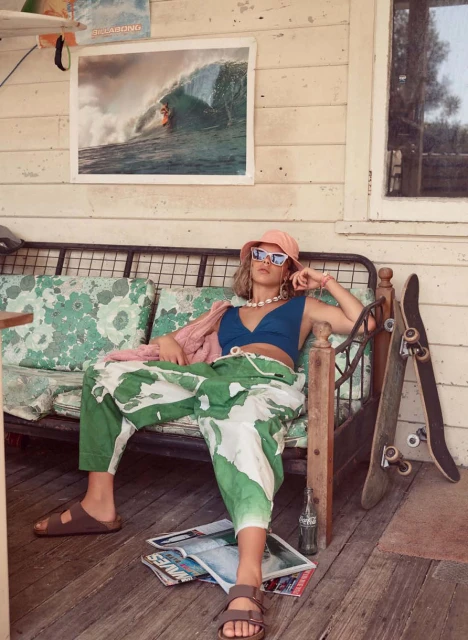 Young woman in bucket hat and stylish clothes lounges on a bench near a skateboard and magazines, creating a relaxed, summery scene.