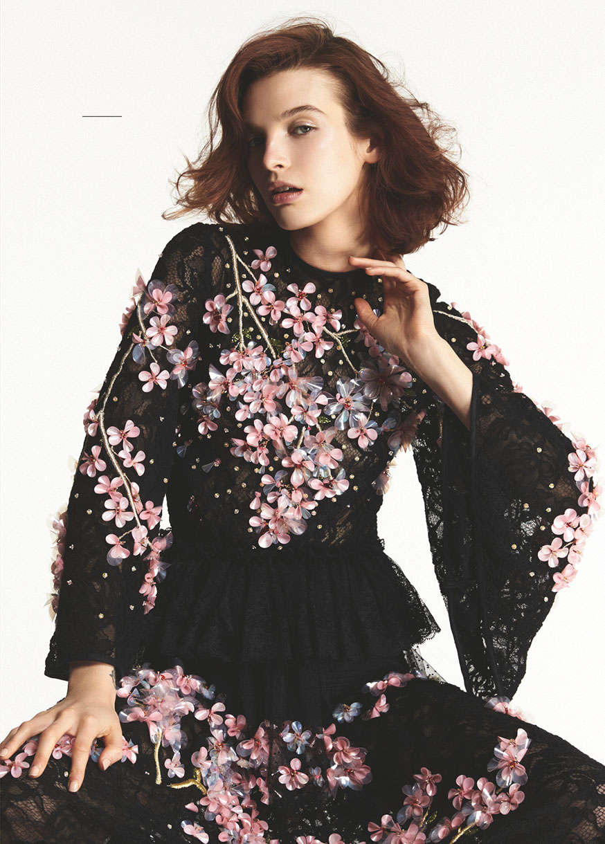 An adult female model poses in a black, long-sleeved, floral-embellished lace dress with flared sleeves against a white backdrop.