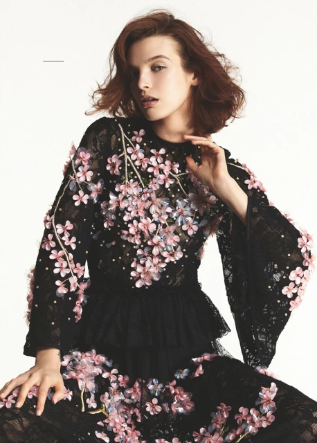 An adult female model poses in a black, long-sleeved, floral-embellished lace dress with flared sleeves against a white backdrop.