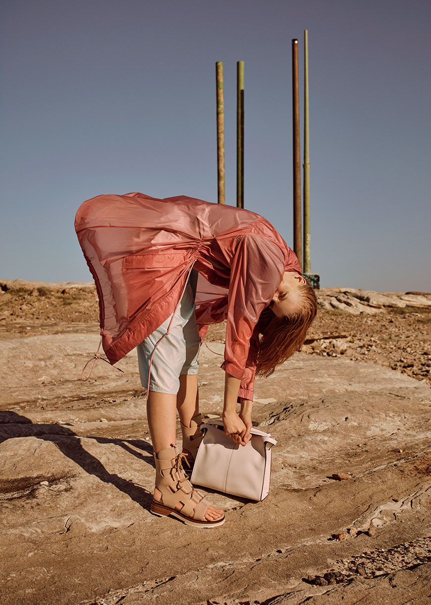 Woman in a sheer pink jacket and gladiator sandals bends over holding a light pink handbag against a rocky outdoor setting.