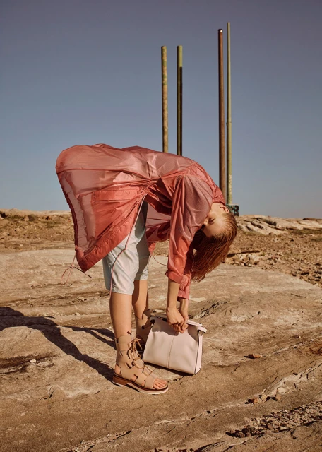 Woman in a sheer pink jacket and gladiator sandals bends over holding a light pink handbag against a rocky outdoor setting.