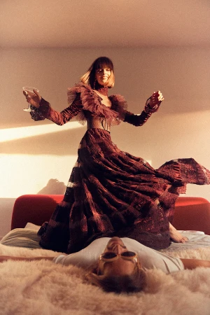 Woman in elaborate gown poses with drink above reclining person on shag rug in warm, natural light, high-fashion, surreal scene.