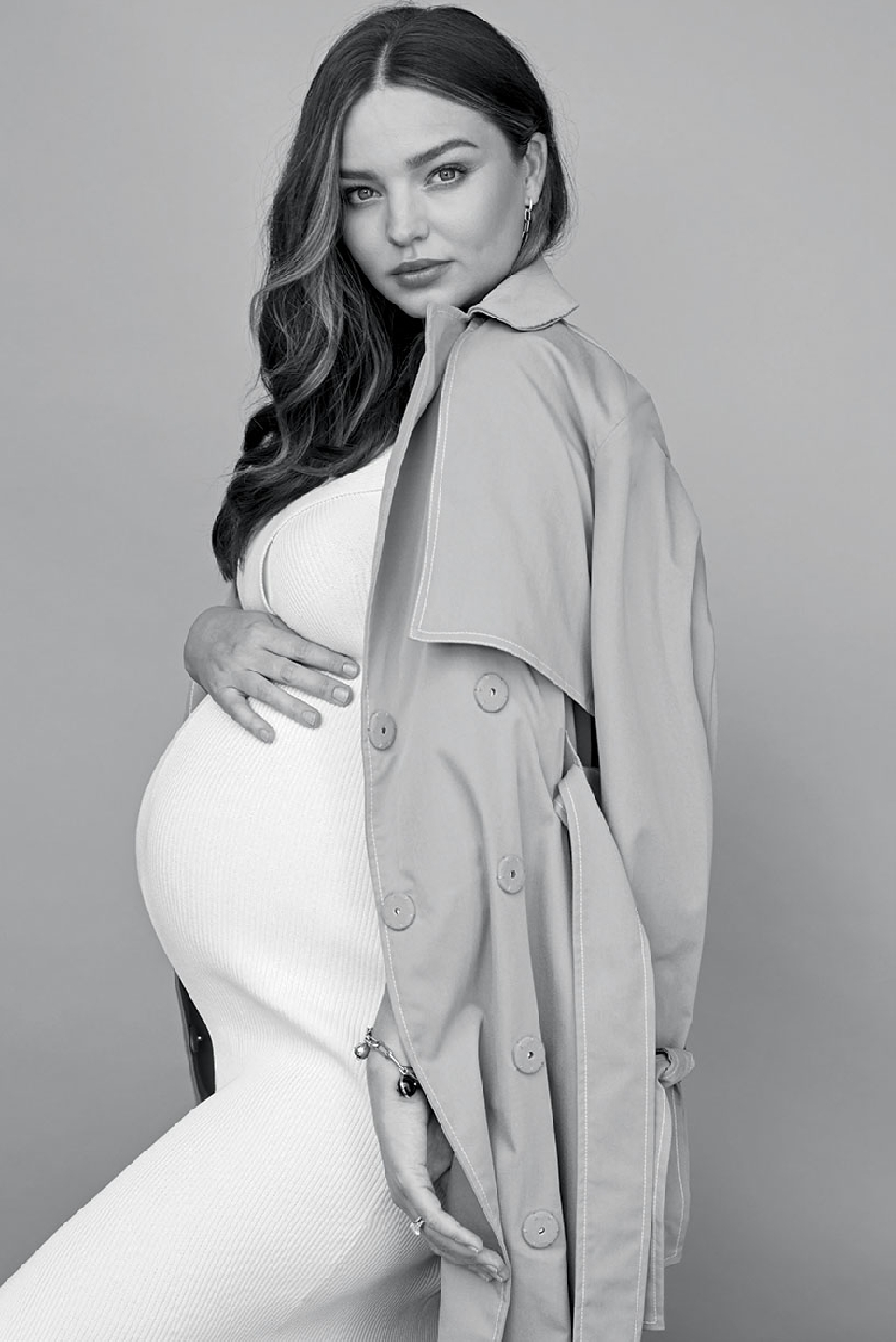 Black and white portrait of a pregnant woman in a white dress, wearing a trench coat draped over her shoulders against a neutral backdrop.