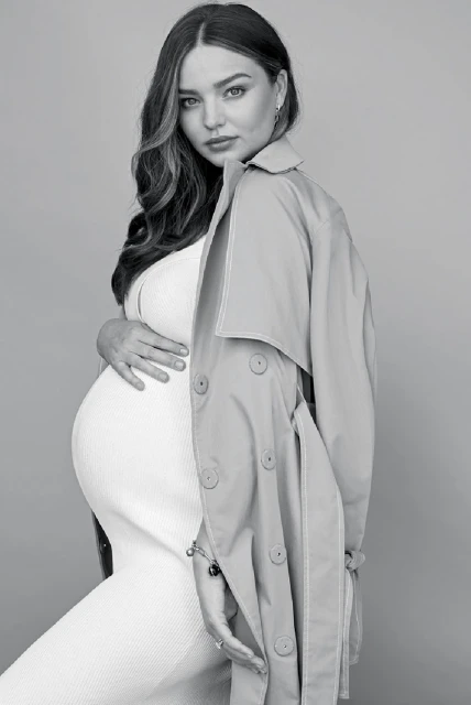Black and white portrait of a pregnant woman in a white dress, wearing a trench coat draped over her shoulders against a neutral backdrop.