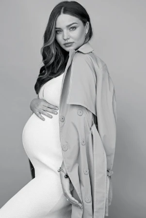 Black and white portrait of a pregnant woman in a white dress, wearing a trench coat draped over her shoulders against a neutral backdrop.