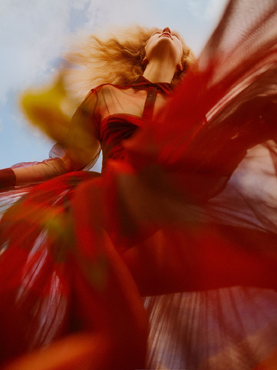 Woman in flowing red sheer dress gazes upward against a blue sky with wispy clouds, caught in a dramatic low-angle fashion shot.
