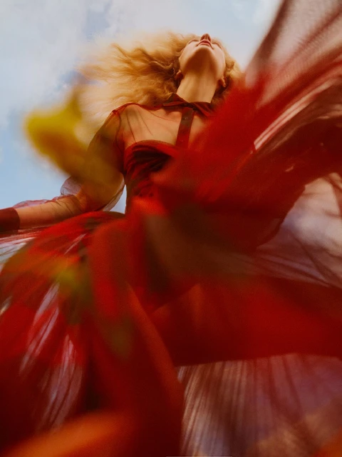 Woman in flowing red sheer dress gazes upward against a blue sky with wispy clouds, caught in a dramatic low-angle fashion shot.