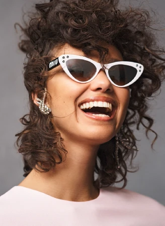 Smiling woman with dark curly hair wearing white cat-eye sunglasses and a silver ear cuff against a grey background