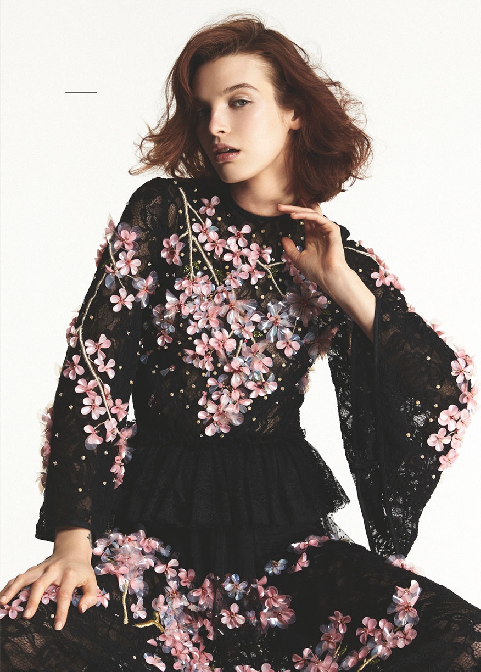Young woman poses in a black lace dress with pink floral embellishments in a studio setting, showcasing fashion and style.