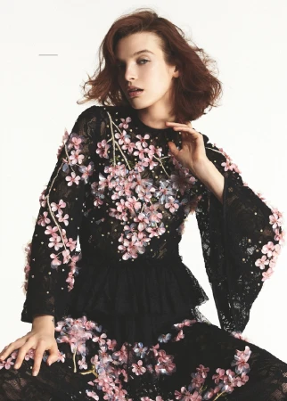 Model with tousled brown hair wearing a black lace dress adorned with pink floral appliqués against a plain white background.