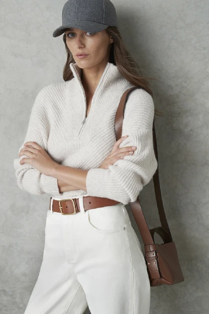 Woman in cream-colored sweater, white jeans, and grey baseball cap with a brown leather purse in a studio setting.