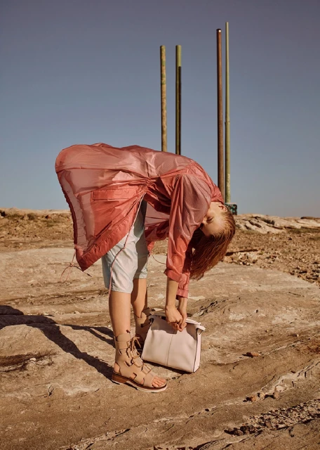 Woman bends over on a rocky surface, wearing a red sheer jacket, holding a light-colored handbag, and wearing gladiator sandals.