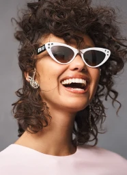 Smiling woman with dark curly hair wearing white cat-eye sunglasses and a silver ear cuff against a grey background