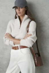 Model wearing a grey baseball cap, white zip-neck sweater, and white trousers with brown belt