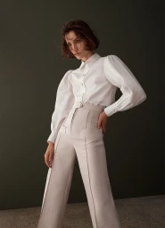 Model with short dark hair wearing a white long-sleeve blouse and high-waist trousers against a dark green backdrop