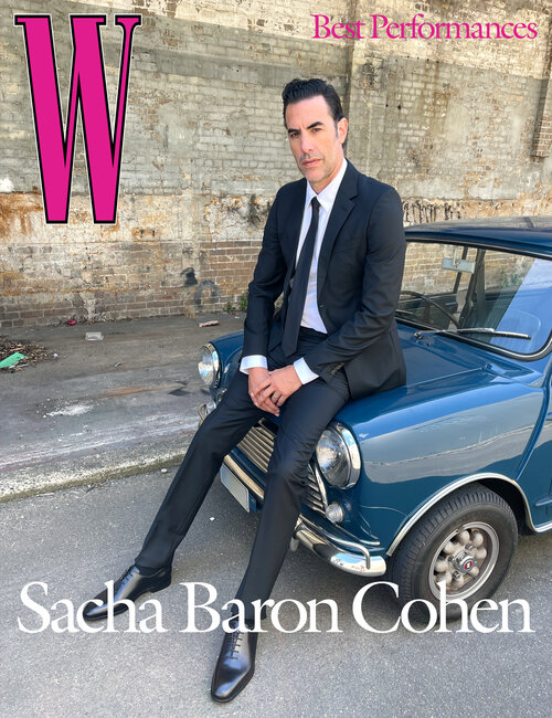 Sacha Baron Cohen sitting on a blue vintage car for a magazine cover with brick wall background and large pink "W" logo.