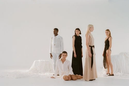 Five diverse fashion models pose in a bright studio setting. One man is seated, wearing a white shirt. The women stand in various dresses.