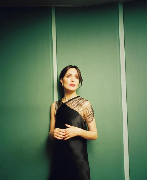 A woman in a black dress poses against a green paneled wall, standing in a three-quarter shot with her hands gently clasped.