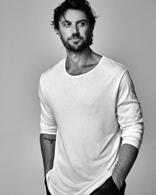 Black and white portrait of a male model in a white long-sleeve shirt, striking a casual pose with his hands in his pockets.
