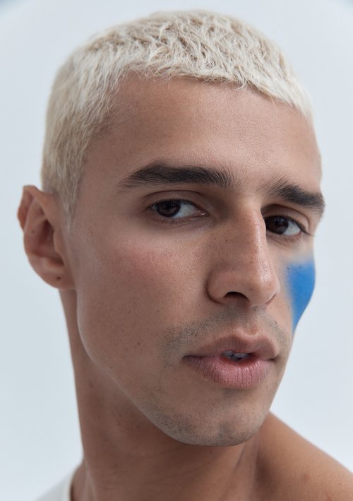 Close-up portrait of a fair-skinned adult male with platinum blonde hair and blue face paint, set against a light grey background.