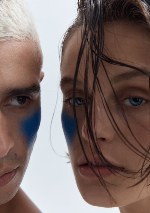 Close-up portrait of two individuals with matching blue face paint, creating a striking contrast against a pale background in a studio setting.