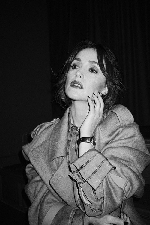 Black and white portrait of a stylish adult woman in a trench coat, looking up with a hand to her face, against a dark background.