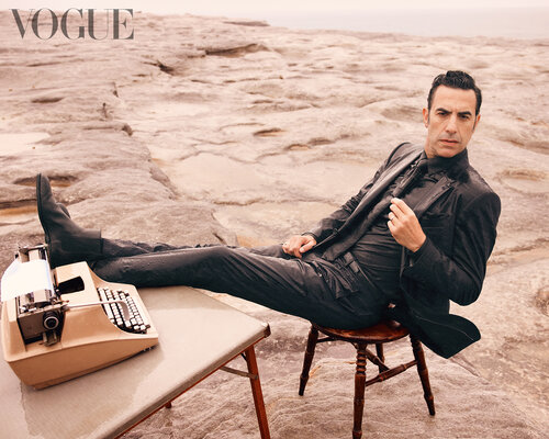 Man in suit lounges next to a vintage typewriter on a rock platform near a beige sand rock formation, overcast outdoor setting.