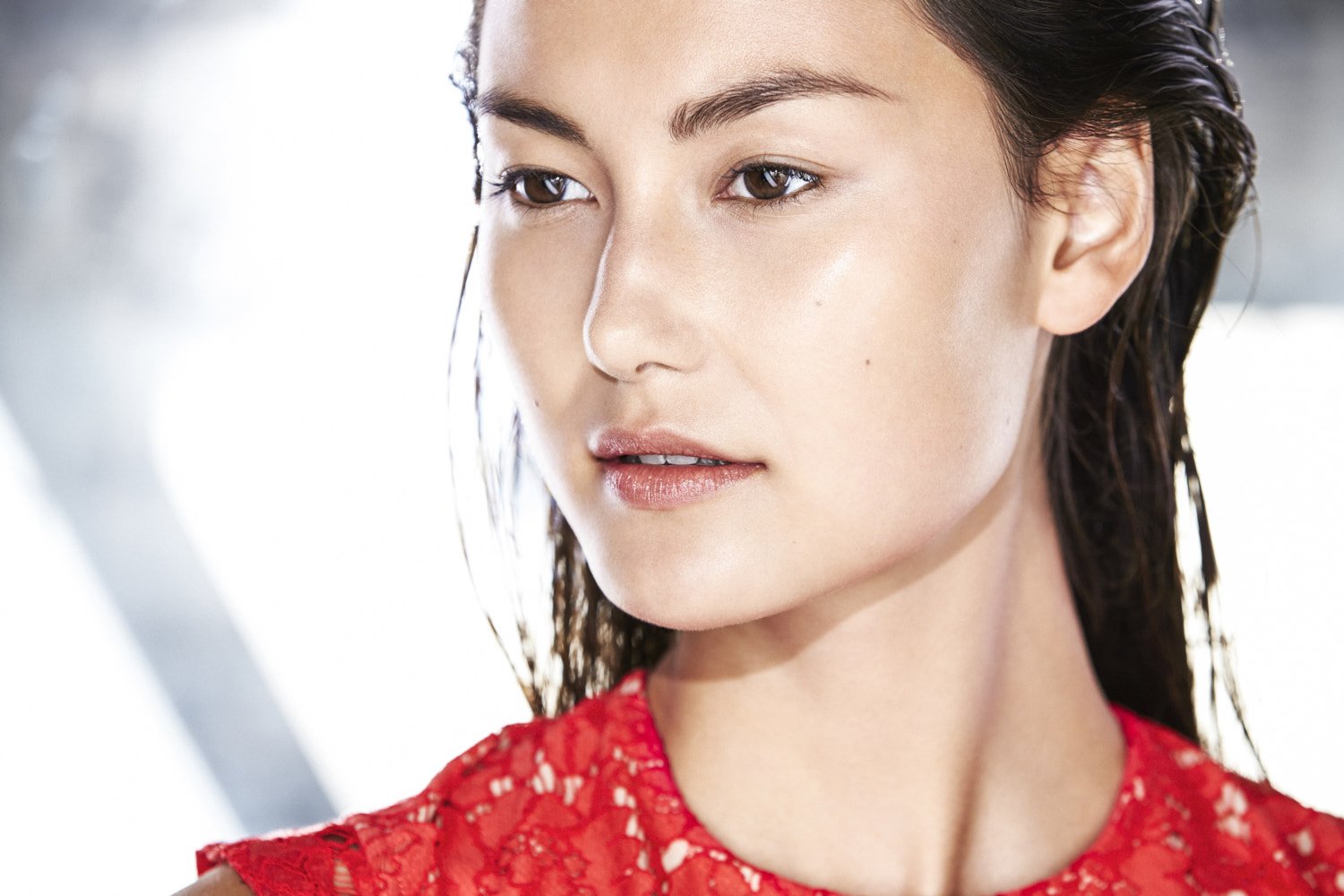 Close-up portrait of a woman with wet hair, wearing a red lace top, showcasing radiant, dewy skin and natural makeup in a bright setting.