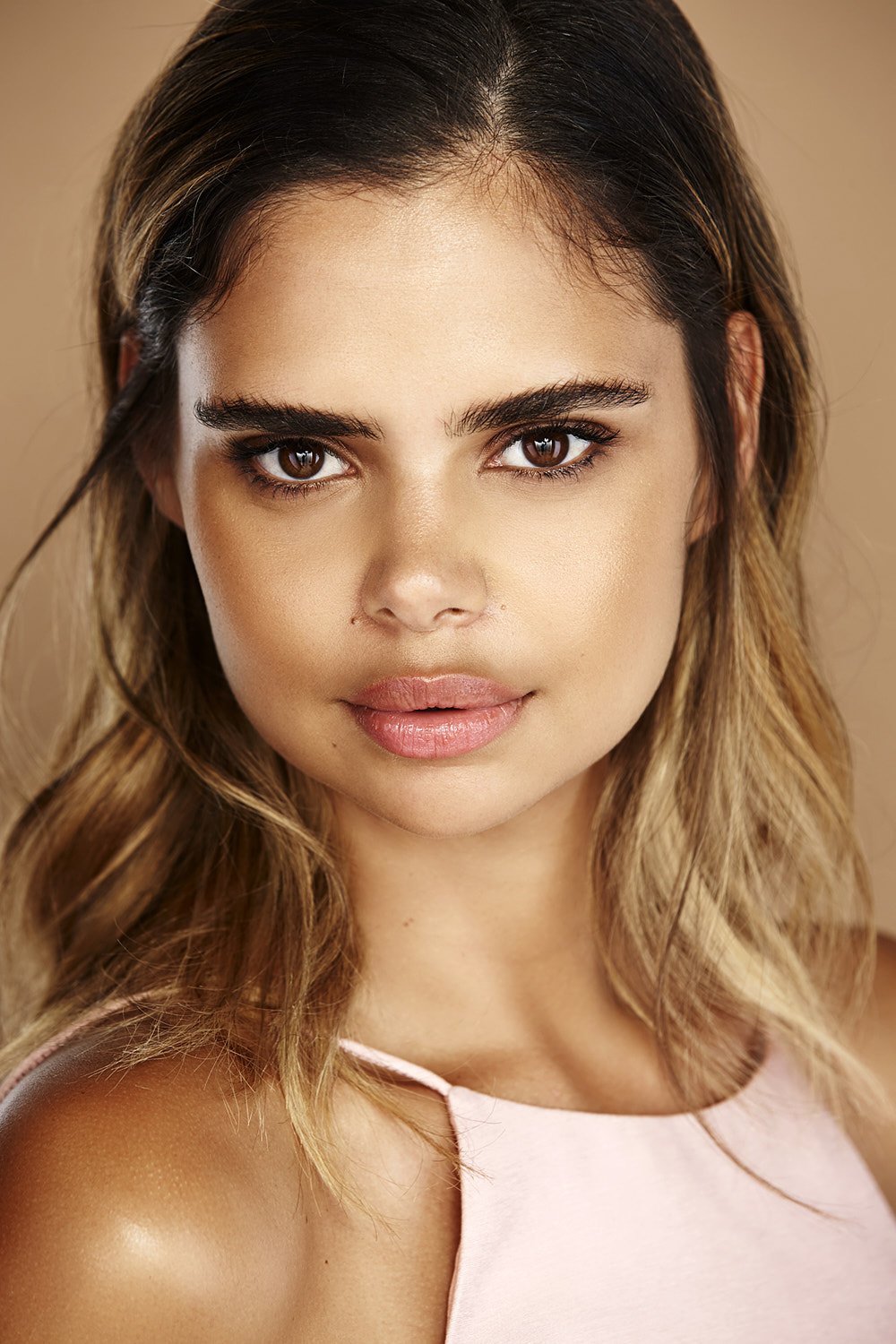 Close-up portrait of a beautiful woman with tan skin, brown eyes, and highlighted brown hair, wearing a pastel pink top against a tan backdrop.