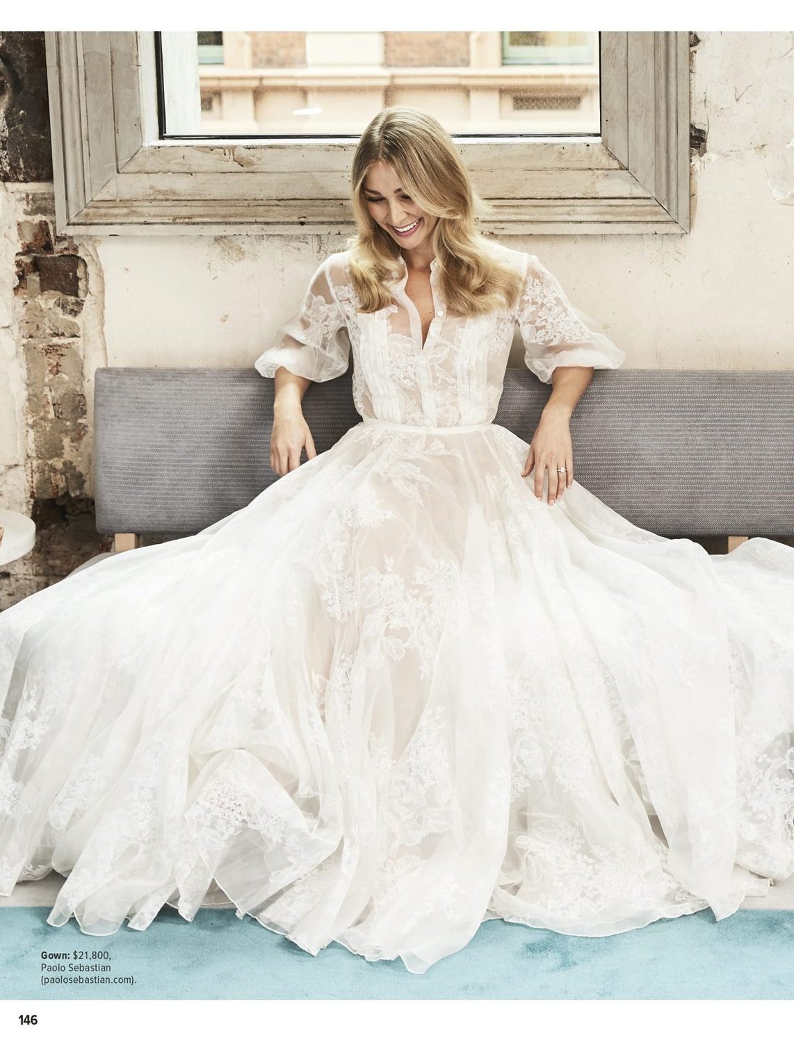 A smiling woman in a lacy white gown sits on a gray bench in front of a window. The background features exposed brick and natural light.