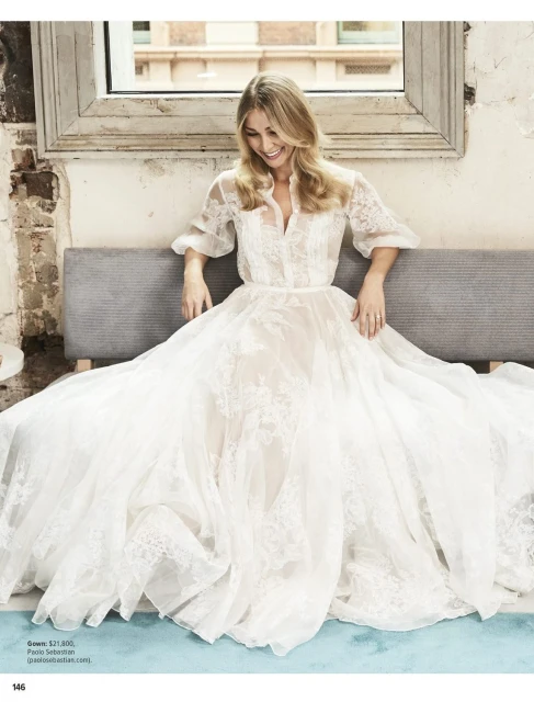 A smiling woman in a lacy white gown sits on a gray bench in front of a window. The background features exposed brick and natural light.