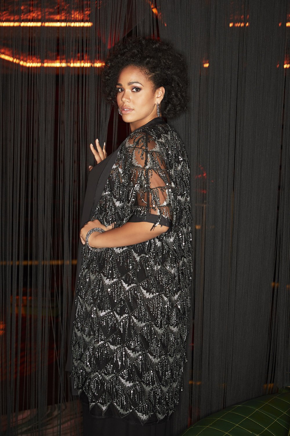 A stylish adult female poses indoors in a black, sequined, sheer kimono-style garment. She has curly dark hair, dark eyes and light skin.