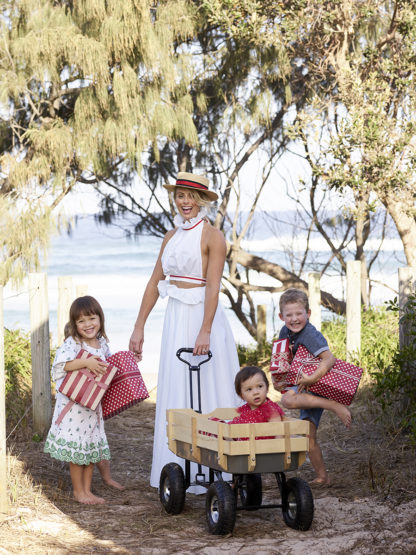 Family of four, including woman, two children, and a baby in a wagon, are posing outdoors with presents near a beach.
