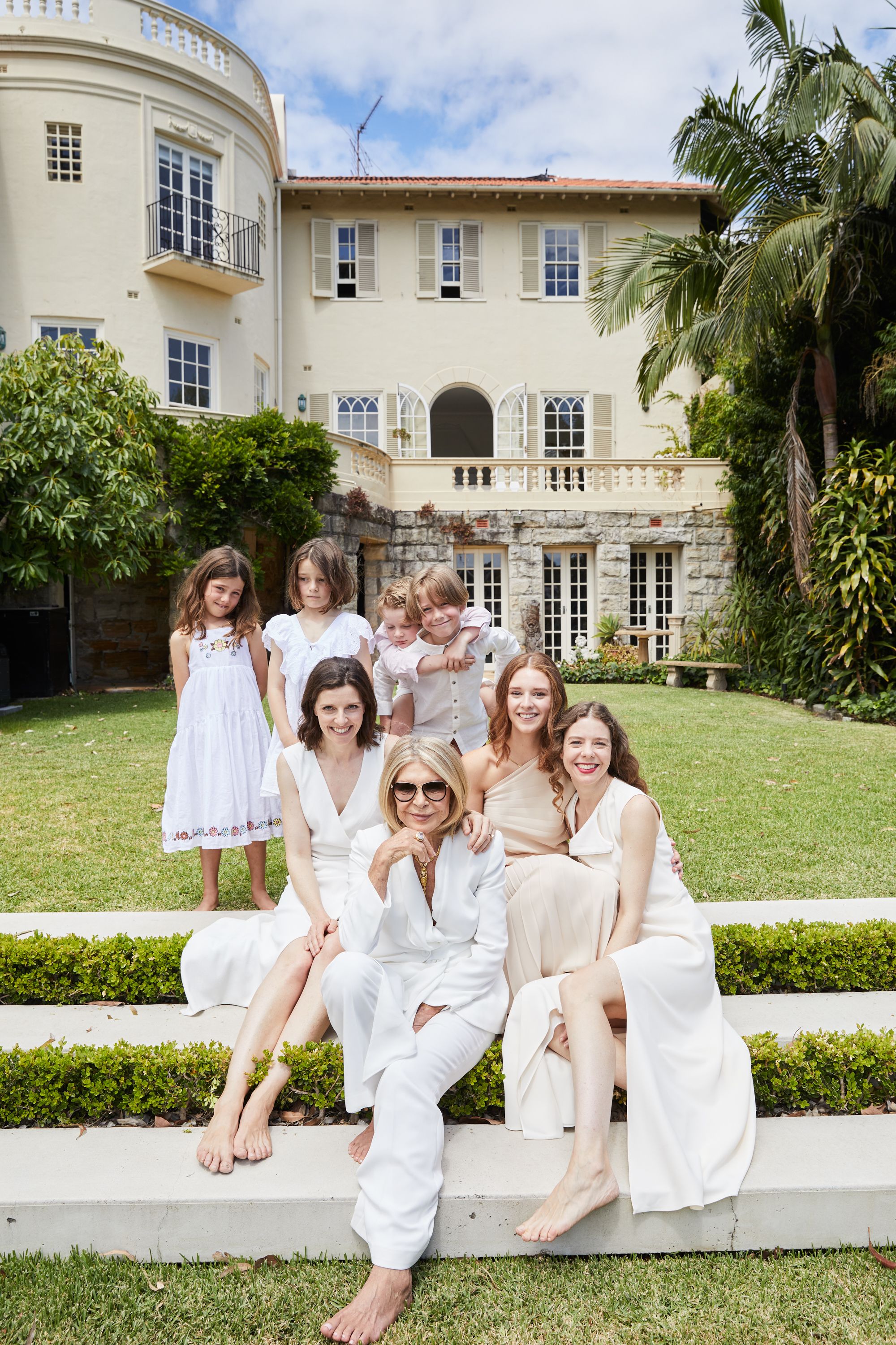 A multigenerational group portrait features three adult women and several children posing on manicured lawn steps in front of a large house.