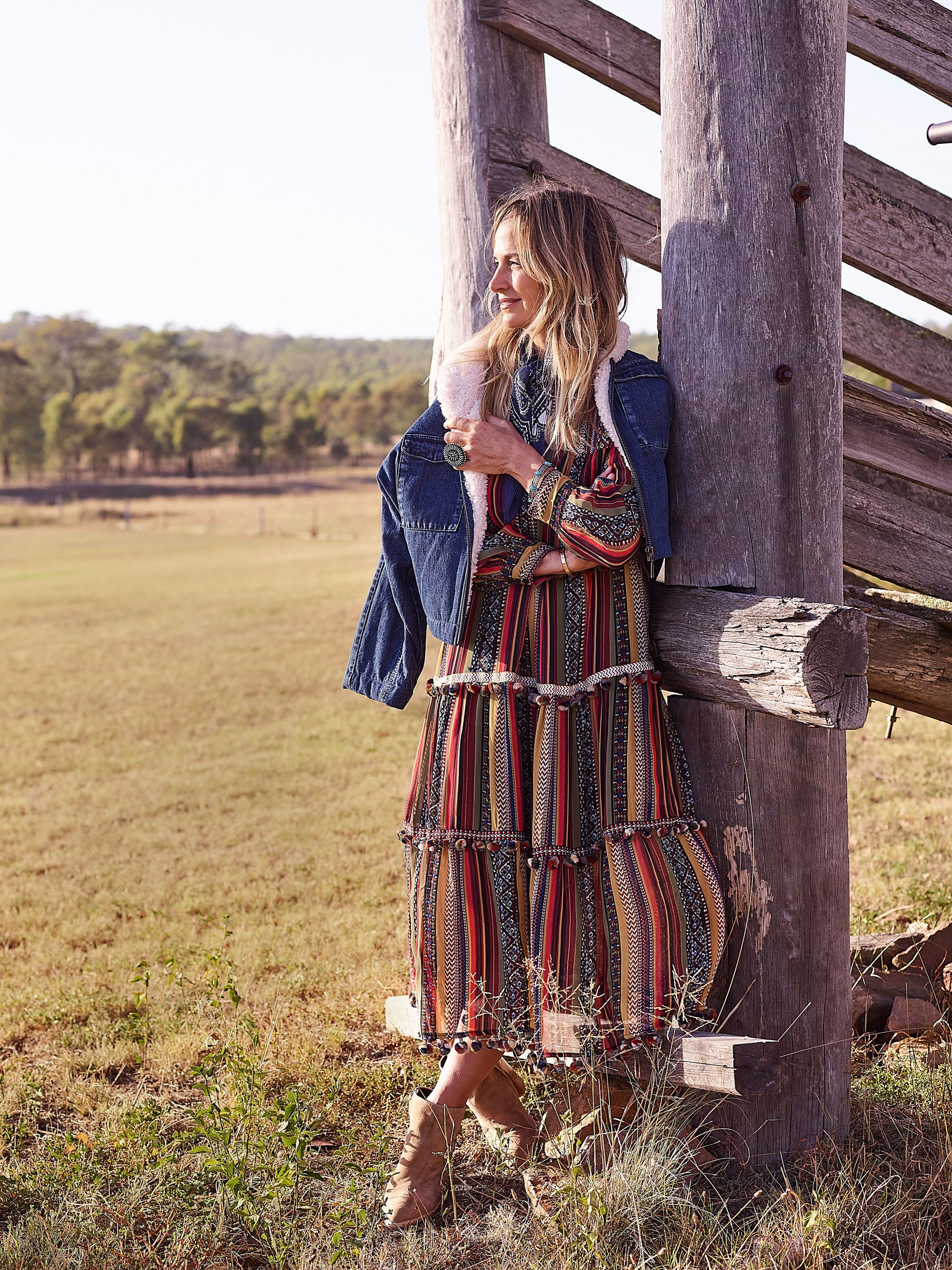Woman in a patterned maxi dress and denim jacket poses near a wooden structure in a sunlit, grassy field with trees in the background.