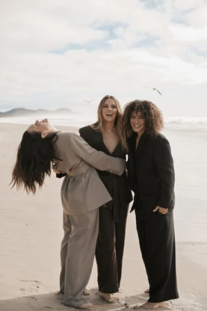 Three women pose in relaxed business attire on a beach with light sand and ocean waves under a bright, cloudy sky.