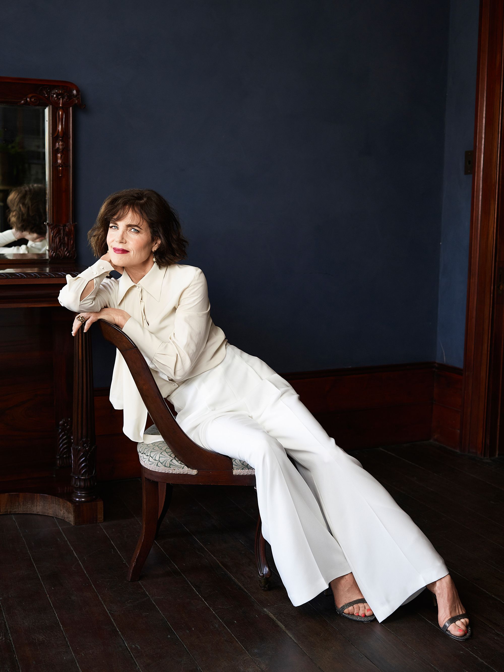 Elegant woman in a cream-colored suit posing while seated on a chair in front of a deep blue wall with a vintage mirror.