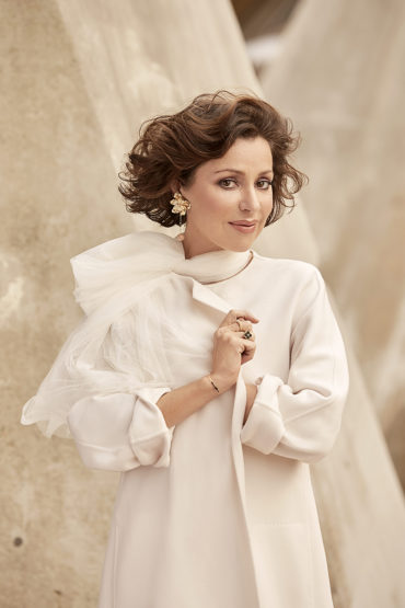 Woman in white coat poses against abstract background. She has short wavy brown hair, a pale complexion, and wears flower earrings.