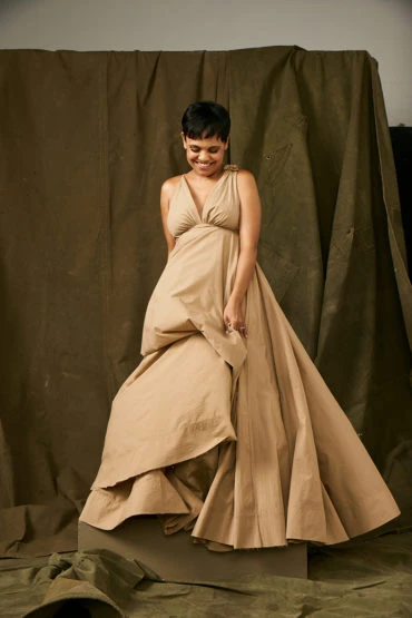Smiling woman in tan gown posing against a draped olive-green backdrop, fashion shoot, studio lighting, simple backdrop, neutral tones.