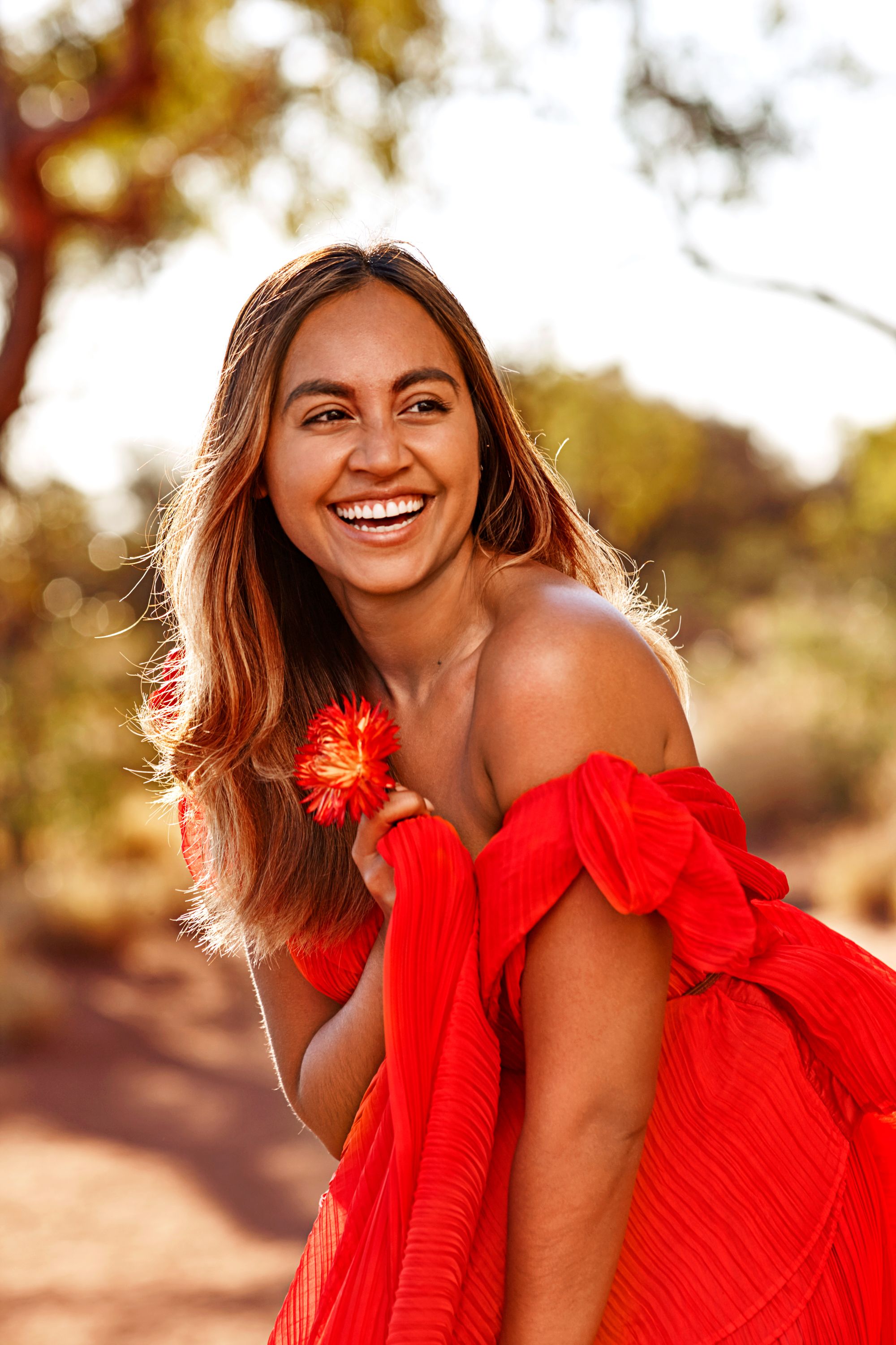 Smiling woman wearing a bright red pleated dress holding a red flower outdoors with trees in the background