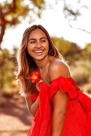 Smiling woman wearing a bright red pleated dress holding a red flower outdoors with trees in the background