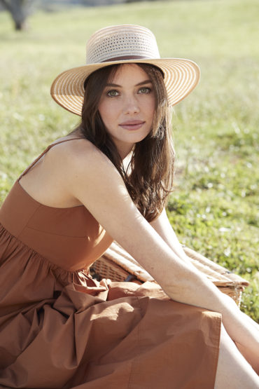 Adult woman wearing a tan sun dress and straw hat, sitting on a blanket with a picnic basket in a grassy outdoor setting.